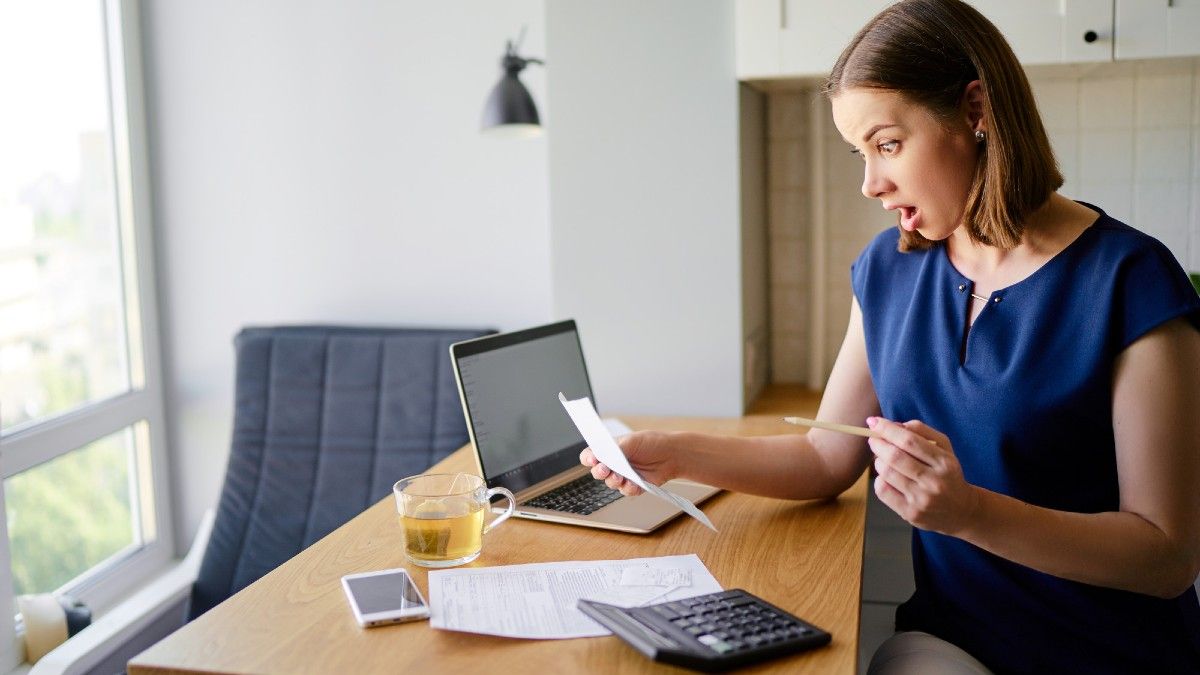 Surprised woman using a laptop computer sitting at her kitchen holding bank statements