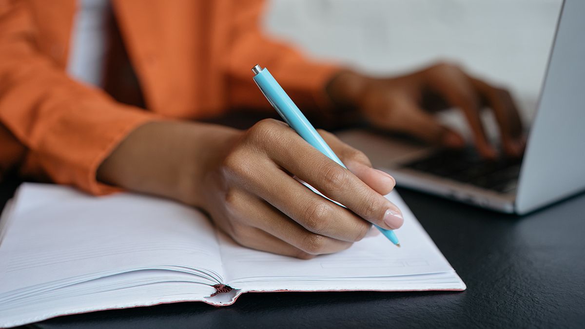 woman holding a pen and writing in a notebook while also using laptop